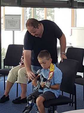 Ein kleiner Junge mit Beinprothese spielt auf seiner Ukulele. Ein Mann beugt sich von hinten zu ihm herab und unterstützt ihn dabei.