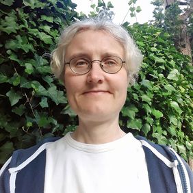 Portraitbild von Elke. Sie hat kurze lockige Haare, schaut freundlich mit geschlossenem Mund direkt in die Kamera und hat eine runde Brille auf.