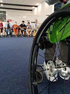Im Vordergrund sieht man ein großes Rollstuhlrad, im Hintergrund ist die Trommelgruppe. Sie besteht aus 5 Jungen, die ca. 12 Jahre als sind.