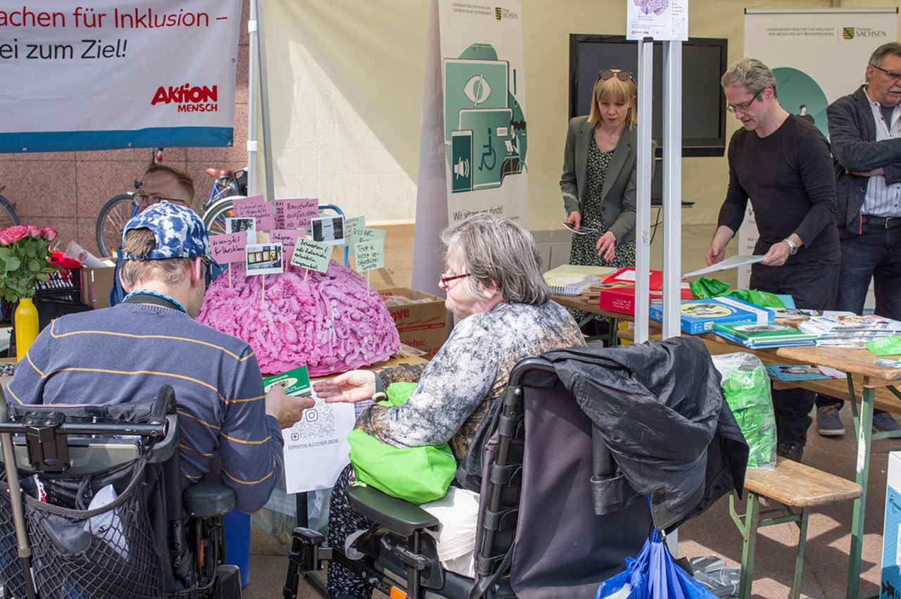 zwei rollstuhlfahrer stehen vor einem Info-Stand mit einem großen Pappmaché-Gehirn