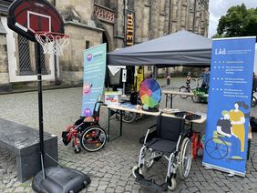 Der Stand der LAG SH Sachsen beim Turnfest in Leipzig. Zu Sehen ist ein Pavillon mit einer Biertischgarnitur auf der ein Glücksrad steht und verschiedene Flyer ausliegen. Vor dem Stand stehen zwei Rollstühle und ein Basketballkorb. Im Hintergrund sieht man die Thomaskirche in Leipzig.