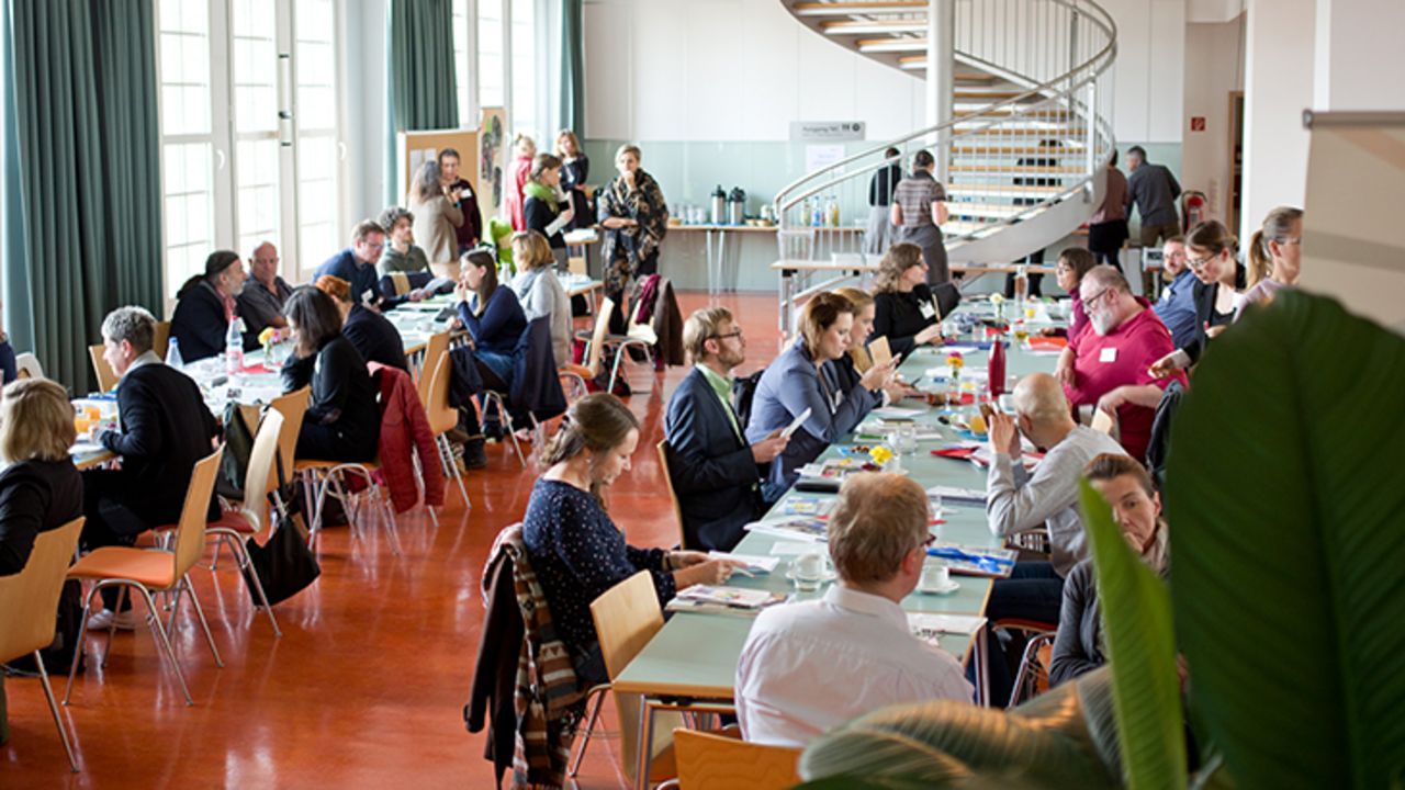 Teilnehmer vom Netzwerktreffen sitzen an langen Tischen, im Hintergrund ist eine Wendeltreppe