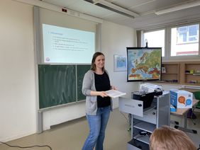 [Translate to Leichte Sprache:] Zu sehen ist Kerstin Helm, die vor einer Tafel steht, während sie einen Vortrag hält. Sie hat braune Haare, lächelt, trägt eine Jeans, ein schwarzes Oberteil und eine graue Strickjacke. Im Hintergrund sieht man eine Schultafel mit einer PowerPoint Präsentation. Rechts im Bild steht eine Landkarte von Europa für den Geographieunterricht und ein Tisch mit technischen Geräten.