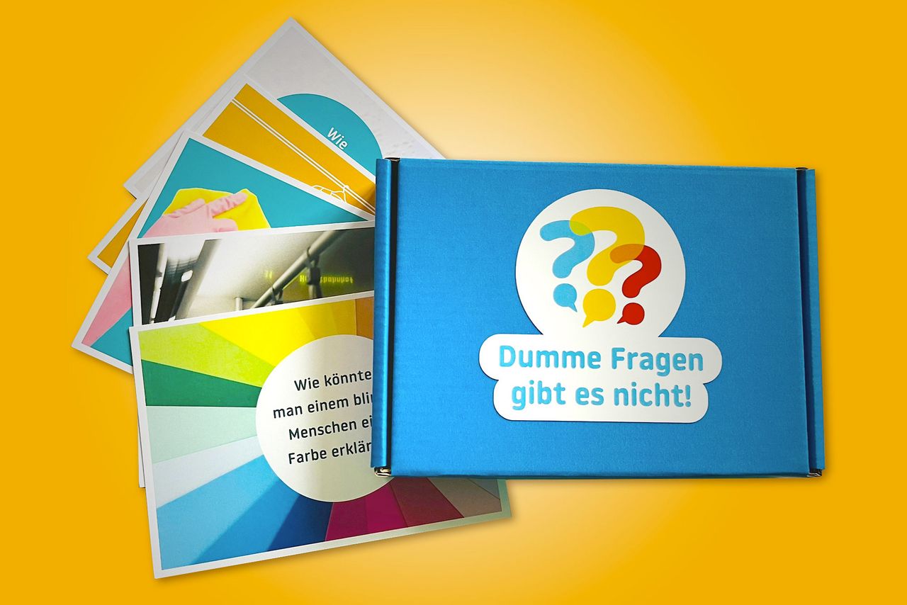 Foto des Kartensets "Dumme Fragen gibt es nicht": einige bunte Karten mit Fragen liegen aufgefächert neben der blauen Box, auf der "Dumme Fragen gibt es nicht" neben drei Fragezeichen steht.