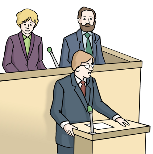 Mann steht am Pult und spricht, dahinter stehen zwei Personen an einem größeren Pult